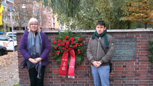 Die Bezirksverordneten Ingrid Bertermann und Leonard Diederich neben dem Gedenkkranz mit roten Blüten der Fraktion DIE LINKE. in der BVV Berlin-Mitte. Auf den Bändern steht der Name der Fraktion und: „Nie wieder – Ihr Schutz ist unsere Pflicht“. 