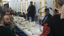 Foto vom Fastenbrechen beim „Haus der Weisheit“. Dieses Bild ist aus der Perspektive von Jacky Sanehy aufgenommen während sie in ein Mikrofon spricht. Zu sehen ist eine große Gruppe Menschen. Einige sitzen an einer Festtafel, andere auf dem Boden. Der Tisch ist reichlich gedeckt.  