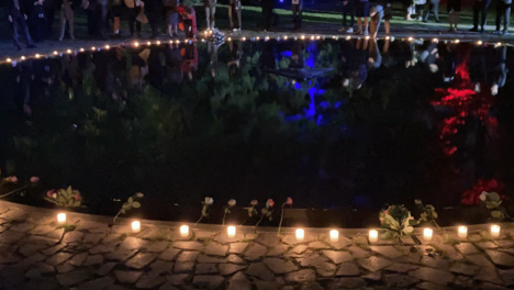 Ein Foto des zentralen Beckens des Mahnmals. Ringsherum wurden Kerzen angezündet und Blumen niedergelegt. Auf der anderen Seite stehen Menschen in Andacht und legen Blumen nieder.