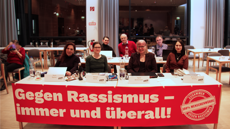 Ein Gruppenfoto der Linken in der BVV Mitte. An dem vorderen Tisch ist ein Banner angebracht auf dem steht „Gegen Rassismus – immer und überall!“. Zu sehen sind in der ersten Reihe von links nach rechts: Deniz Seyhun, Hannah Gerlof, Ingrid Bertermann und Jacky Sanehy. In der zweiten Reihe sitzen von links nach rechts: Samiullah Malekzadeh, Rüdiger Lötzer und Thilo Urchs.