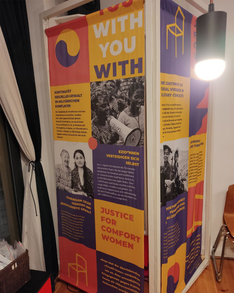 Ein Foto eines Aufstellers im Museum der Trostfrauen zu sehen ist. In Gelb, rot und dunkel Lila sind kurze Artikel über sexualisierte Gewalt in militärischen Konflikte. Schwerpunkt ist der Aktivismus der sich für die Opfer dieser Gewalt einsetzt und sie verhindert. Unten steht groß „Justice for Comfort Women.“