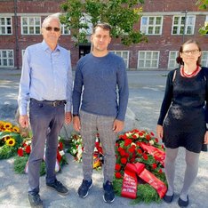 Rüdiger Lötzer, Leonard Diederich und Hannah Gerlof stehen auf dem Rathausvorplatz im Wedding neben einem Kranz mit roten Nelken und einer roten Schleife, der vor einem Gedenkstein liegt. Auf der Schleife steht: Wo Unrecht zu Recht wird, wird Widerstand zur Pflicht. DIE LINKE in der BVV Mitte.