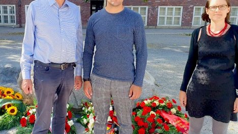 Rüdiger Lötzer, Leonard Diederich und Hannah Gerlof stehen auf dem Rathausvorplatz im Wedding neben einem Kranz mit roten Nelken und einer roten Schleife, der vor einem Gedenkstein liegt. Auf der Schleife steht: Wo Unrecht zu Recht wird, wird Widerstand zur Pflicht. DIE LINKE in der BVV Mitte.