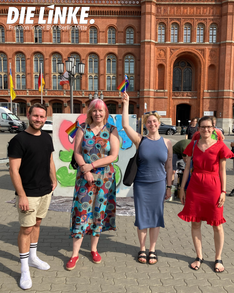 Ein Gruppenfoto vor dem Roten Rathaus. Von Links nach rechts: Christoph Keller, Ingrid Bertermann, Martha Kleedörfer und Hannah Gerlof. Martha hält eine Pride-Fahne. Oben links ist das Logo der Fraktion DIE LINKE. in der BVV Berlin Mitte zu sehen.