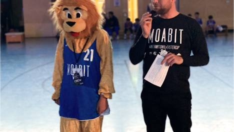 easter cup eröffnung Foto von der Eröffnung des International Easter Cup Berlin Moabit 2024. Rechts zu sehen ist Jugendstadtrat Christoph Keller (Linke) beim Halten seiner Eröffnungsrede. Neben ihm steht eine Person in einem Löwenkostüm in einem Moabit 21 Trikot – das Maskottchen des Easter Cups.