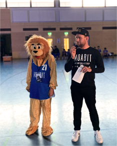 easter cup eröffnung Foto von der Eröffnung des International Easter Cup Berlin Moabit 2024. Rechts zu sehen ist Jugendstadtrat Christoph Keller (Linke) beim Halten seiner Eröffnungsrede. Neben ihm steht eine Person in einem Löwenkostüm in einem Moabit 21 Trikot – das Maskottchen des Easter Cups.