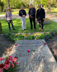 Gruppenfoto: Die Bezirksverordneten Sven Diedrich Martha Kleedörfer und Thilo Urchs sowie eine weitere Person stehen vor dem Gedenkstein in der Seestraße. Auf dem steht: „Hier ruhen 295 Opfer der Nationalsozialistischen Diktatur“. Rote Rosen und ein Blumenkranz aus roten Rosen und anderen roten Blüten wurden auf dem Stein abgelegt, dafür blühen in einem kleinem Beet Frühlingsblumen.