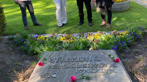 Gruppenfoto: Die Bezirksverordneten Sven Diedrich Martha Kleedörfer und Thilo Urchs sowie eine weitere Person stehen vor dem Gedenkstein in der Seestraße. Auf dem steht: „Hier ruhen 295 Opfer der Nationalsozialistischen Diktatur“. Rote Rosen und ein Blumenkranz aus roten Rosen und anderen roten Blüten wurden auf dem Stein abgelegt, dafür blühen in einem kleinem Beet Frühlingsblumen.