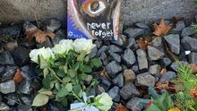 Foto der Gaben die beim Gleis 69 in Moabit niedergelegt wurden. Zu sehen sind weiße Rosen der Omas gegen Rechts und ein Kunstwerk, das ein schwarzen Hamsa zeigt mit der Inschrift „Never Forget“. 