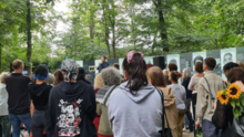 Foto vom Protest gegen die Entfernung des Mahnmals an den Porajmos in Berlin Tiergarten. Es ist eine große Gruppe Menschen zu sehen, die einem Redebeitrag vor den Gedenktafeln lauscht.