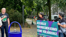 Foto der Bezirksverordneten Ingrid Bertermann während ihres Redebeitrages bei der Kundgebung an der Friedensstatue in Berlin-Moabit. Neben ihr stehen zwei ältere Frauen mit einem Banner auf dem steht „Sexuelle Verbrechen kennen keine Verjährung“. 