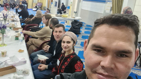 Foto vom Fastenbrechen beim „Haus der Weisheit“. Im Vordergrund sind Samiullah Haidary, Jacky Sanehy und Leonard Diederich zu sehen – Bezirksverordnete für die Fraktion DIE LINKE. in der BVV Mitte. Im Hintergrund sind weitere Menschen an einer Tafel zu sehen sowie mehrere Menschen, die auf einer Bühne sitzen. Der Raum ist festlich eingerichtet. 