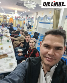 Foto vom Fastenbrechen beim „Haus der Weisheit“. Im Vordergrund sind Samiullah Haidary, Jacky Sanehy und Leonard Diederich zu sehen – Bezirksverordnete für die Fraktion DIE LINKE. in der BVV Mitte. Im Hintergrund sind weitere Menschen an einer Tafel zu sehen sowie mehrere Menschen, die auf einer Bühne sitzen. Der Raum ist festlich eingerichtet. 