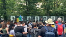 Foto vom Protest gegen die Entfernung des Mahnmals an den Porajmos in Berlin Tiergarten. Es ist eine große Gruppe Menschen mit Fahnen und Plakaten zu sehen, die einem Redebeitrag vor den Gedenktafeln lauscht.