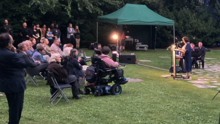 Foto von der Gedenkveranstaltung für den Porajmos am 2. August beim Mahnmal Tiergarten. Rechts steht eine Person die einen Redebeitrag hält. Links sitzen und stehen Menschen während sie den Redebeitrag zuhören.