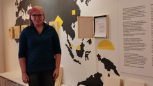 Foto der Bezirksverordneten Ingrid Bertermann. Sie guckt in die Kamera. Im Hintergrund ist ein Teil der Ausstellung sichtbar – Bleistiftportraits auf Holz die an einer Wand mit einer Weltkarte angebracht sind. Außerdem ist die Überschrift eines Ausstellungstextes erkennbar, dort steht „Wer waren die Trostfrauen?“